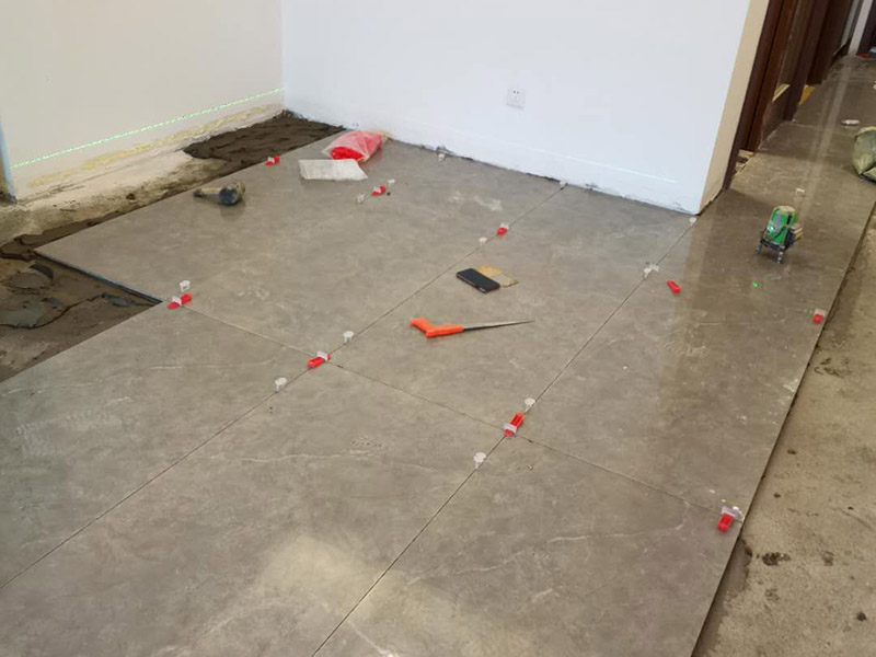 客廳地磚鋪貼：地磚鋪貼前，應(yīng)將地面清掃干凈，尤其是凝結(jié)后的混凝土，否則，地磚鋪貼后會(huì)出現(xiàn)翹邊、空鼓等現(xiàn)象，然后再灑水潤(rùn)濕地面。地面充分濕潤(rùn)，但不能有積水，然后將3:1混合好的沙子水泥鋪在地面上，進(jìn)行基層處理。在地磚背面均勻抹水泥砂漿作為粘合層，厚度不低于1cm，然后鋪在剛才試鋪的位置上，用橡皮錘輕敲地磚飾面直至密實(shí)平整達(dá)到要求。地磚鋪完后24h進(jìn)行清理勾縫，勾縫前應(yīng)先將地磚縫隙內(nèi)雜質(zhì)擦凈，用專(zhuān)用填縫劑勾縫，完工后（一般宜在24h之后）再用棉紗等物對(duì)地磚表面進(jìn)行清理。